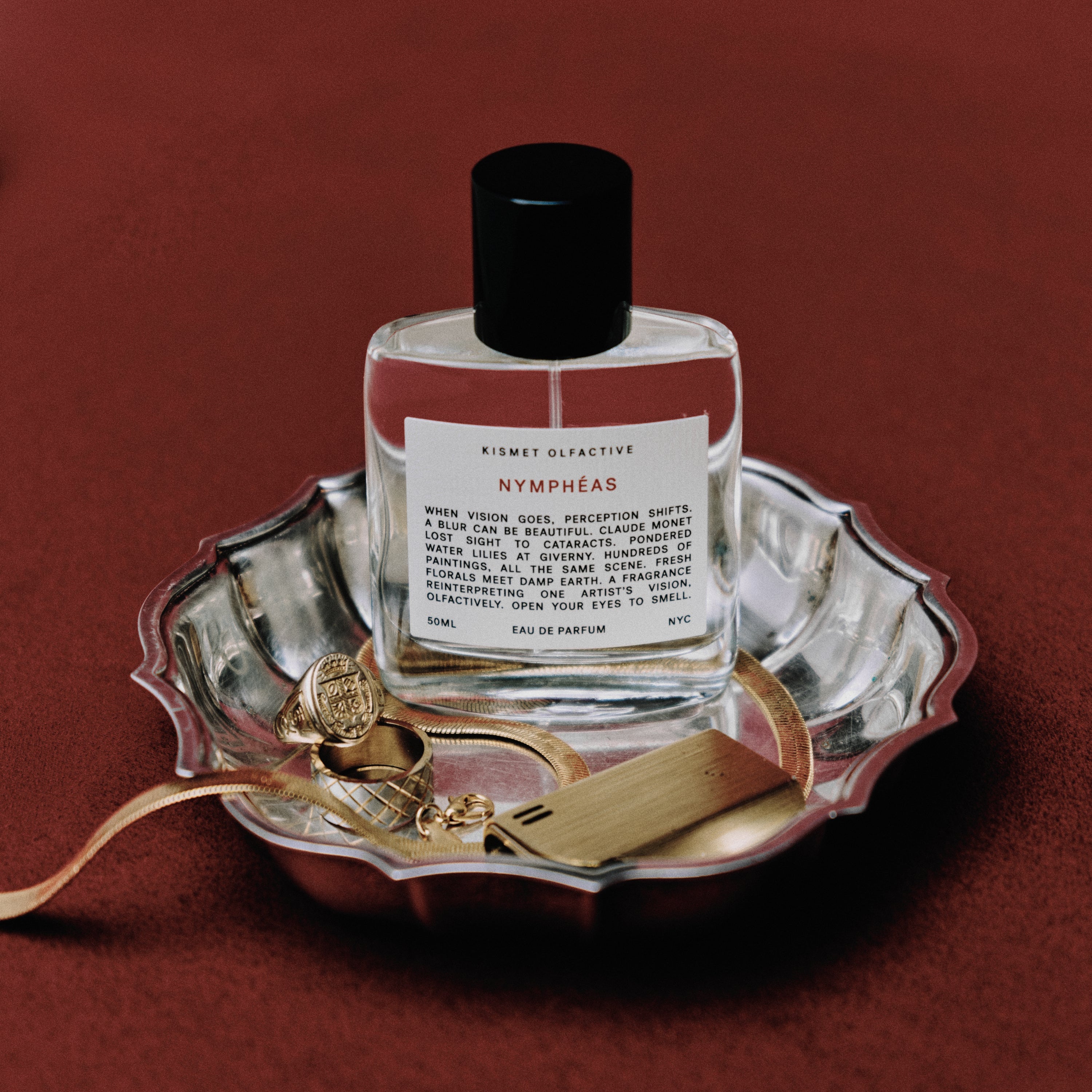 Perfume bottle by Kismet Olfactive labeled 'Nymphéas' on a decorative tray with gold accents against a red background